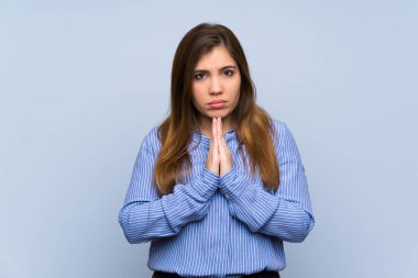 Young girl over isolated blue wall keeps palm together. Person asks for something