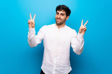 Genç adam mavi duvarın üstünde gülümsüyor ve iki eliyle zafer işareti gösteriyor.