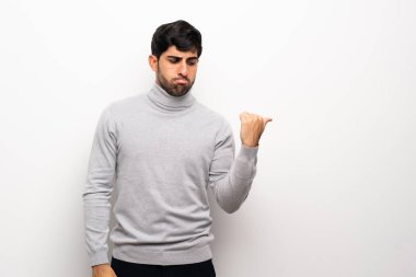 Genç adam izole edilmiş beyaz duvarın üzerinde mutsuz ve kenarı işaret ediyor.