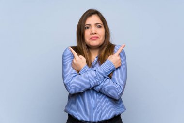 Young girl over isolated blue wall pointing to the laterals having doubts