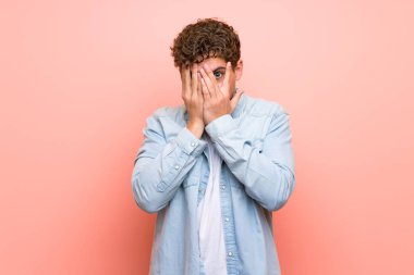 Elleri ile gözleri kaplayan ve parmaklardan bakarak pembe duvar üzerinde Blonde adam