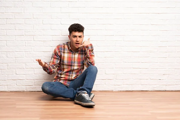 Mladý Muž Sedící Podlaze Který Dělá Telefonní Gesto Pochybuje — Stock fotografie