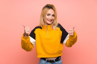 Soyutlanmış pembe bir geçmişi olan genç sarışın bir kadın. Baş parmakları havada ve gülümsüyor.