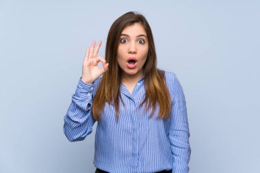 Young girl over isolated blue wall surprised and showing ok sign
