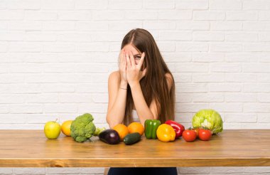 Bir sürü sebzesi olan genç bir kadın gözlerini kapatıyor ve parmaklarından bakıyor.