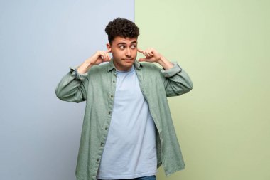 Mavi ve yeşil geçmişi olan genç bir adam iki kulağını da elleriyle kapatıyor.