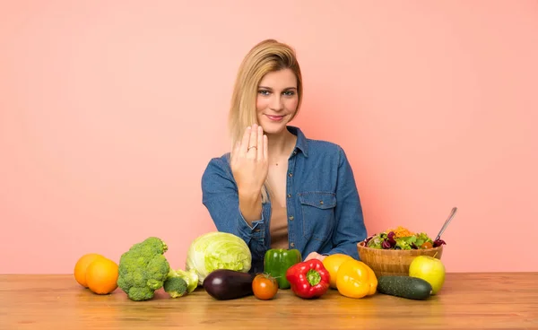 Young blonde woman with many vegetables inviting to come with hand. Happy that you came