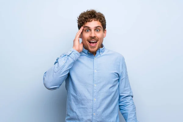 Blonde man over blue wall with surprise and shocked facial expression