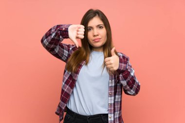 Young girl over pink wall making good-bad sign. Undecided between yes or not