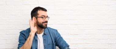 Beyaz tuğla duvar üzerinde sakallı yakışıklı adam kulağına el koyarak bir şey dinlerken