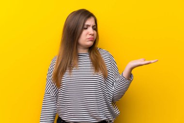 Young girl over yellow wall holding copyspace with doubts