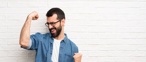 Handsome man with beard over white brick wall celebrating a victory