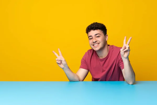 Renkli Duvarı Masası Olan Genç Adam Iki Eliyle Zafer Işareti — Stok fotoğraf