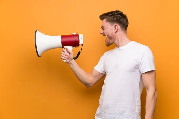 Redhead man over brown wall shouting through a megaphone