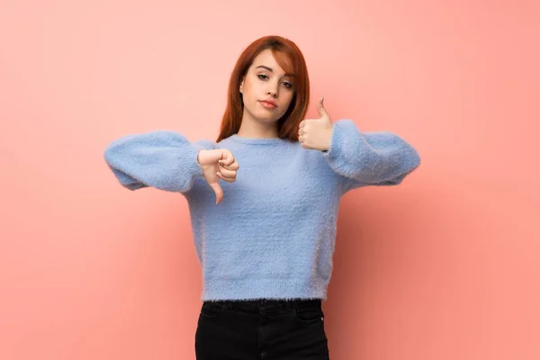 Young redhead woman over pink background making good-bad sign. Undecided between yes or not