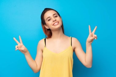 Gülümseyen ve iki elini zafer işaretiyle gösterilen mavi arka plan genç Kızıl saçlı kadın