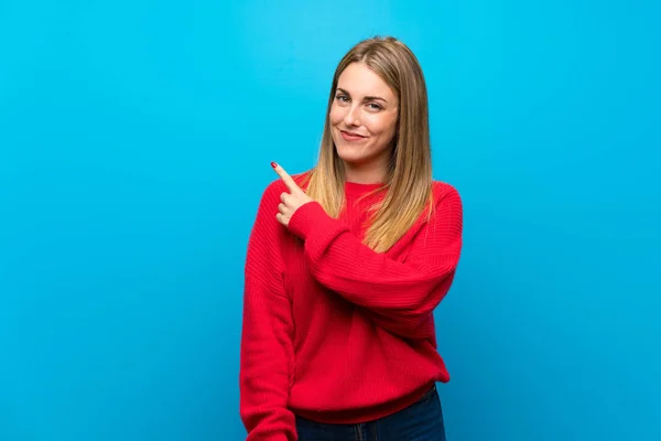 Woman with red sweater over blue wall pointing to the side to present a product