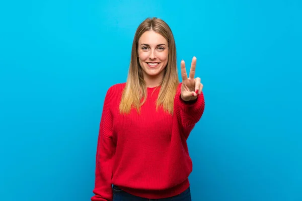 Woman with red sweater over blue wall smiling and showing victory sign