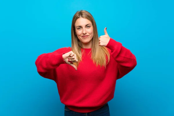 Woman with red sweater over blue wall making good-bad sign. Undecided between yes or not