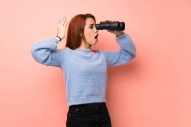 Pembe bir arka plan ve Uzaktan dürbünle bakarak genç Kızıl saçlı kadın