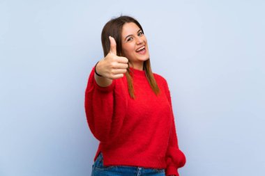 Genç bir kadın, mavi duvardan izole edilmiş. Başparmakları havada. Çünkü iyi bir şey oldu.