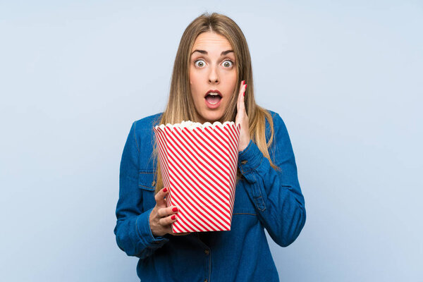 Blonde woman with popcorns over blue wall doing surprise gesture