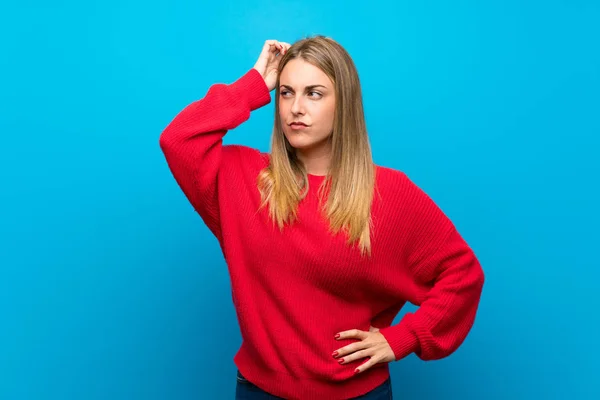 Woman with red sweater over blue wall having doubts while scratching head