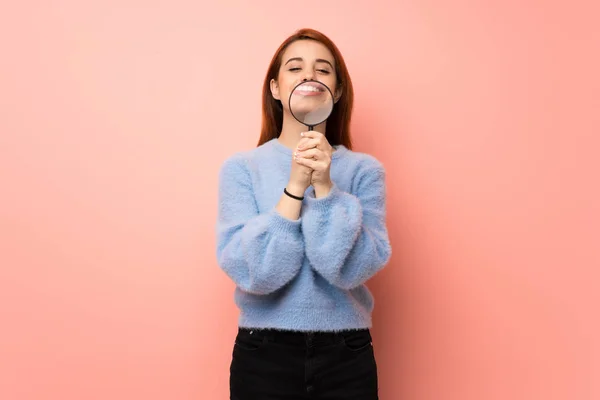 Young redhead woman over pink background taking a magnifying glass and showing teeth through it