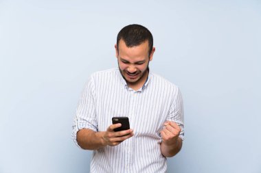 Zafer pozisyonunda telefon ile izole mavi duvar üzerinde Kolombiyalı adam