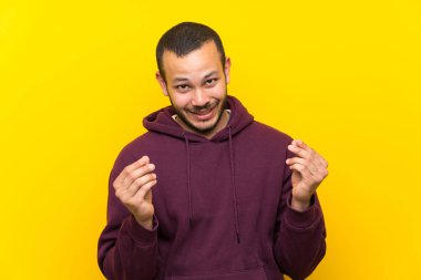Sarı duvar üzerinde sweatshirt ile Kolombiyalı adam para jest yapma