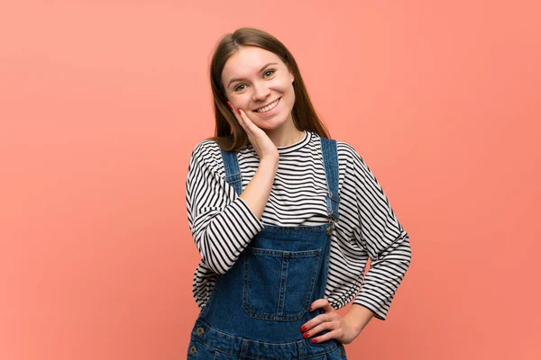 Young woman with overalls over pink wall smiling and looking to the front with confident face