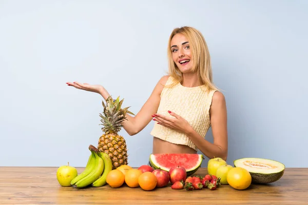 Young blonde woman with lots of fruits extending hands to the side for inviting to come