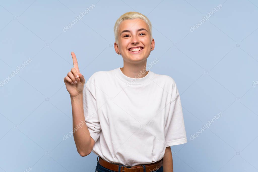 Teenager girl with white short hair over blue wall showing and lifting a finger in sign of the best