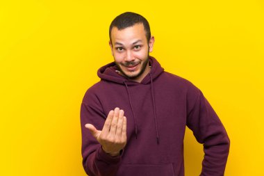 Sarı duvar üzerinde sweatshirt ile Kolombiyalı adam el ile gelmeye davet ediyor. Geldiğin için mutluyum.