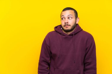 Sarı duvar üzerinde sweatshirt ile Kolombiyalı adam yan görünümlü şüpheler jest yapma