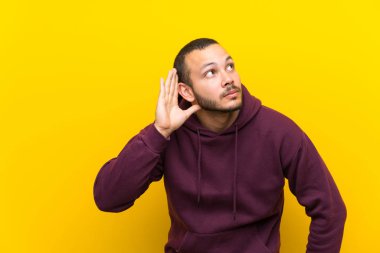 Sarı duvar üzerinde Sweatshirt ile Kolombiyalı adam kulak el koyarak bir şey dinliyor