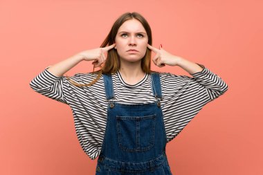 Pembe duvarda tulumlu genç bir kadın iki kulağını da elleriyle kapatıyor.