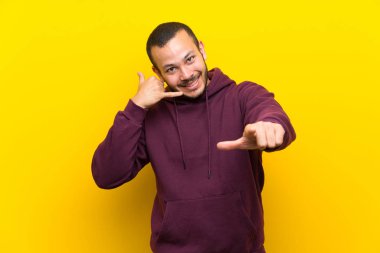 Sarı duvar üzerinde sweatshirt ile Kolombiyalı adam telefon jest yapma ve ön işaret