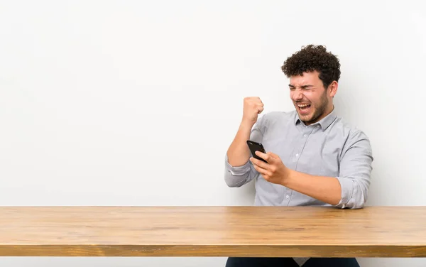 Junger Mann Mit Tisch Und Telefon Siegerpose — Stockfoto