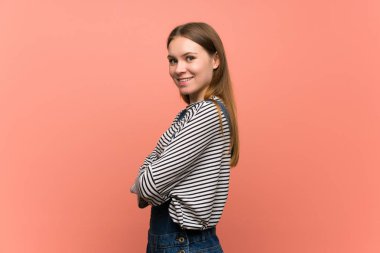 Pembe duvarda tulumlu genç bir kadın gülümseyerek kollarını yan yana tutuyor.