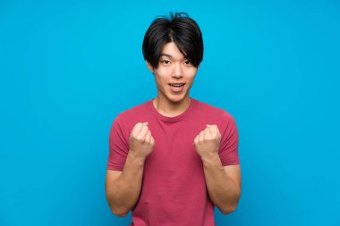 İzole mavi duvar üzerinde kırmızı gömlekli Asyalı adam zaferi kutluyor