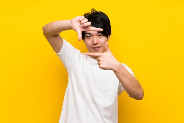 Asian man over isolated yellow wall focusing face. Framing symbol