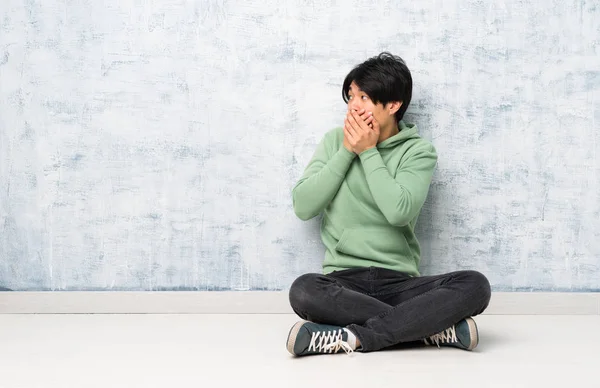 Asian man sitting on the floor covering mouth and looking to the side