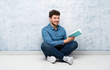 Genç adam yerde oturmuş bir kitap tutuyor ve okuyor.