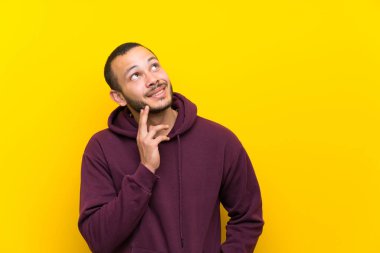 Sarı duvar üzerinde sweatshirt ile Kolombiyalı adam ararken bir fikir düşünme