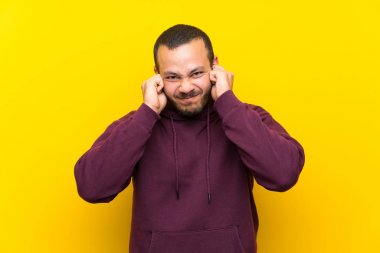 Sarı duvar üzerinde sweatshirt ile Kolombiyalı adam sinirli ve kulakları kapsayan