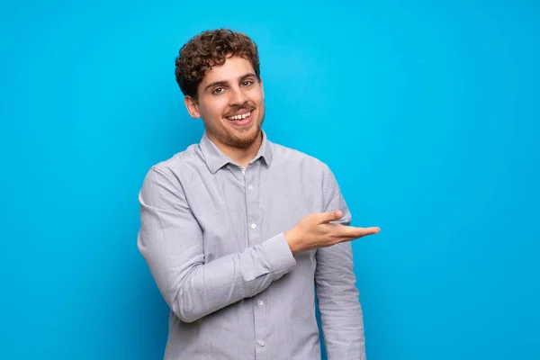 Blonde man over blue wall presenting an idea while looking smiling towards