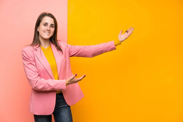 Young Woman Pink Suit Colorful Background Extending Hands Side Inviting — Zdjęcie stockowe
