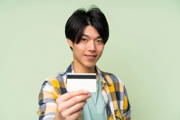 Asian man over isolated green wall holding a credit card