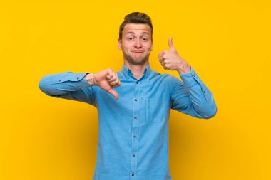 Izole sarı duvarın üstündeki sarışın adam iyi-kötü işareti yapıyor. Evet ya da değil arasında kararsız